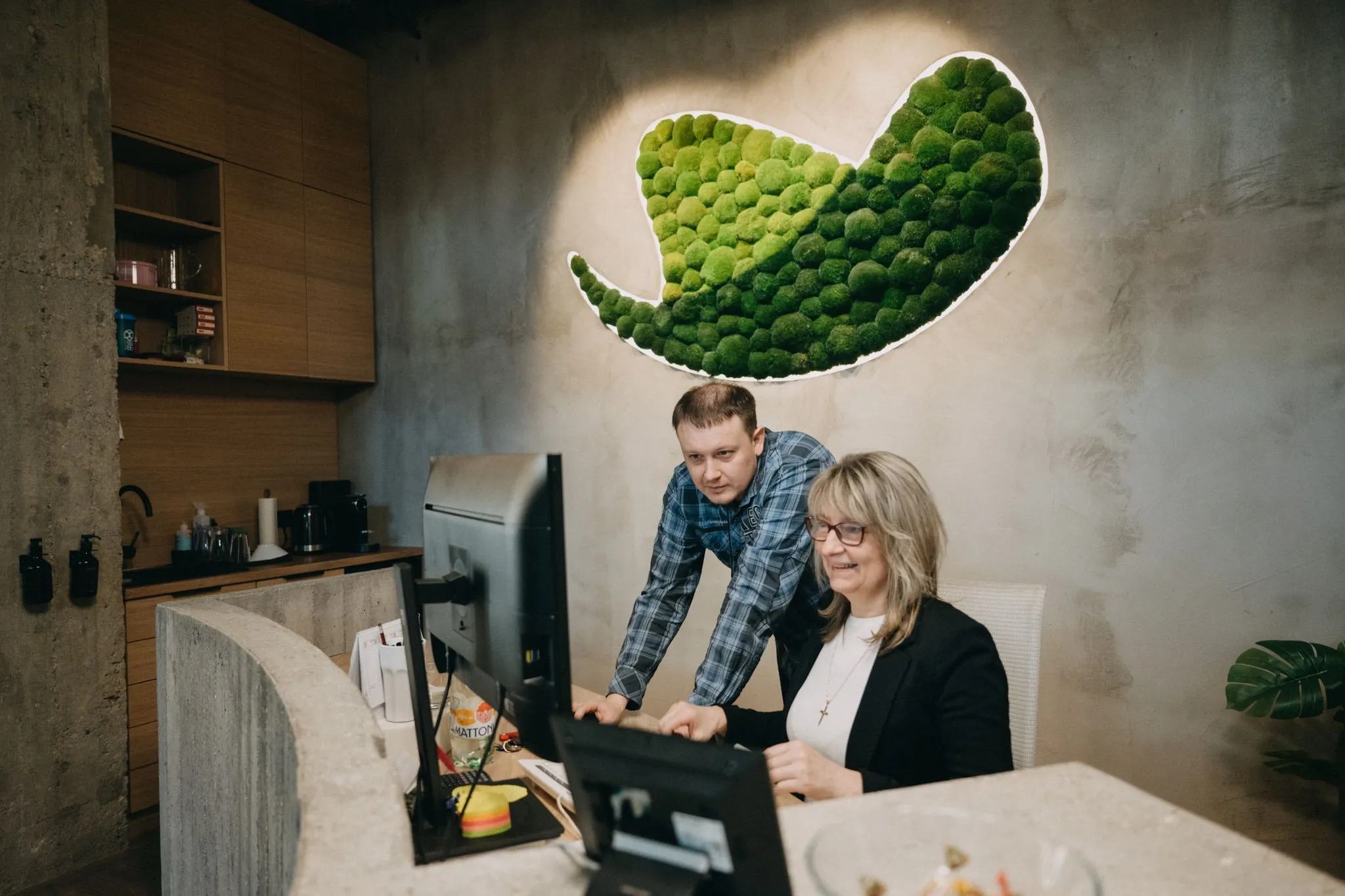 Two colleagues collaboratively working at a computer desk with unique green moss wall art.