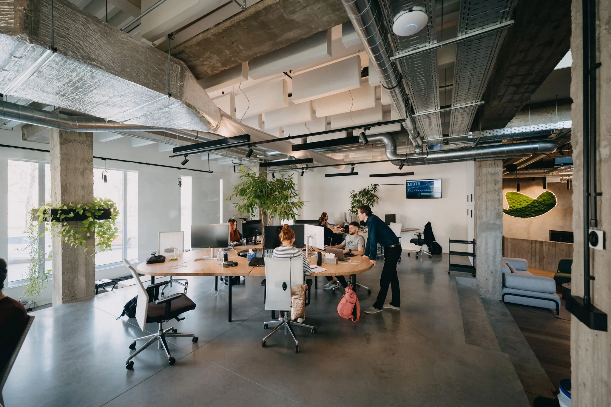 Modern open-plan office with employees working at desks and lush green plants.
