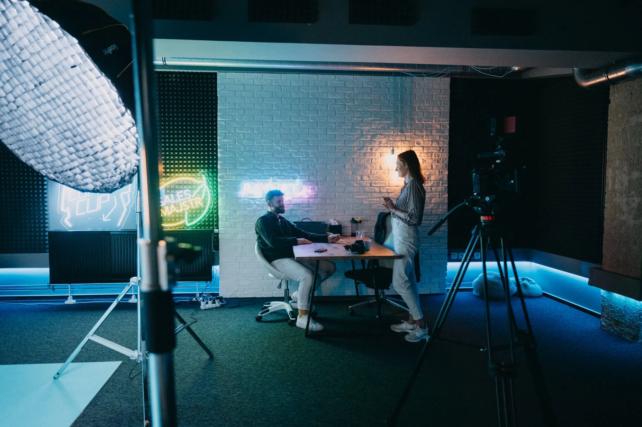 Two people work at a table in a neon-lit studio with filming equipment and blue lighting.