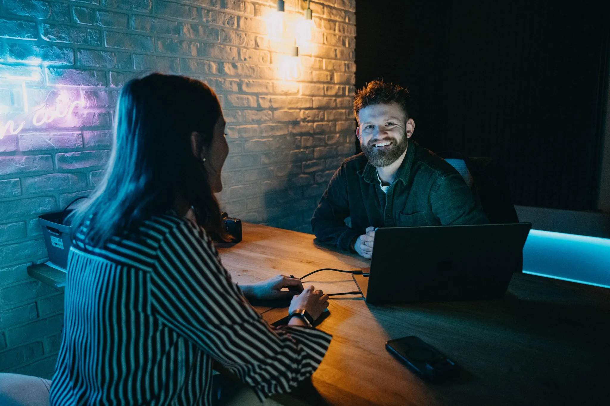 Man smiling at woman across a table with a laptop in a dimly lit room.