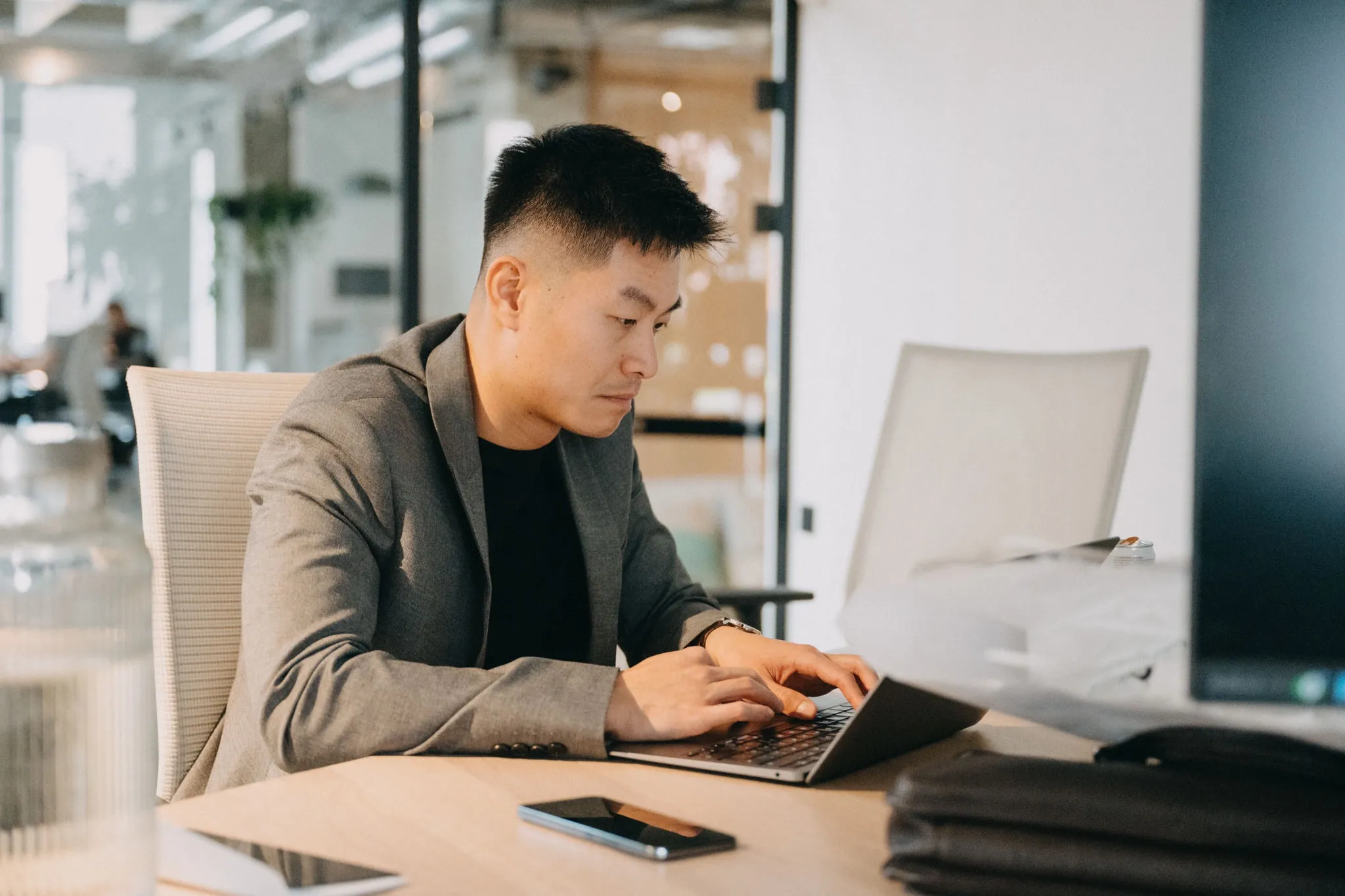 Young Asian man in a suit typing intently on a laptop at a modern office desk.