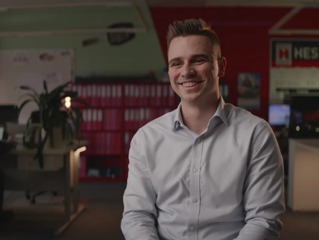 A smiling young man in a light shirt sits in an office setting.