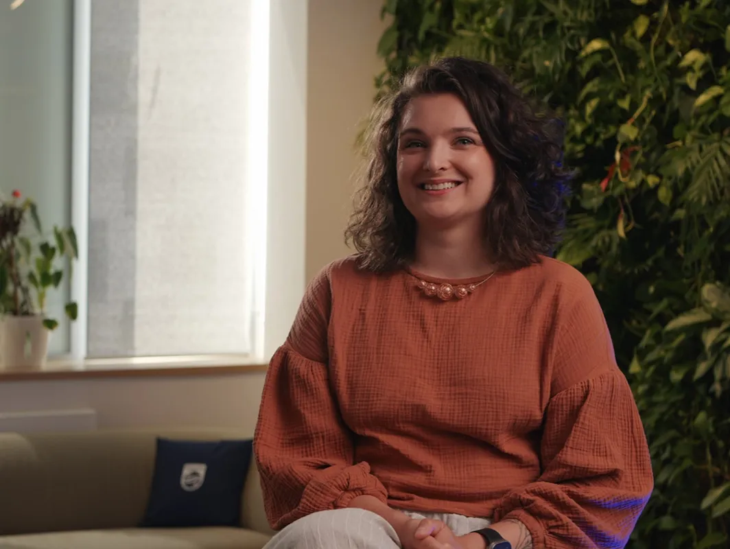 A smiling woman with curly hair sits indoors against a green plant wall.