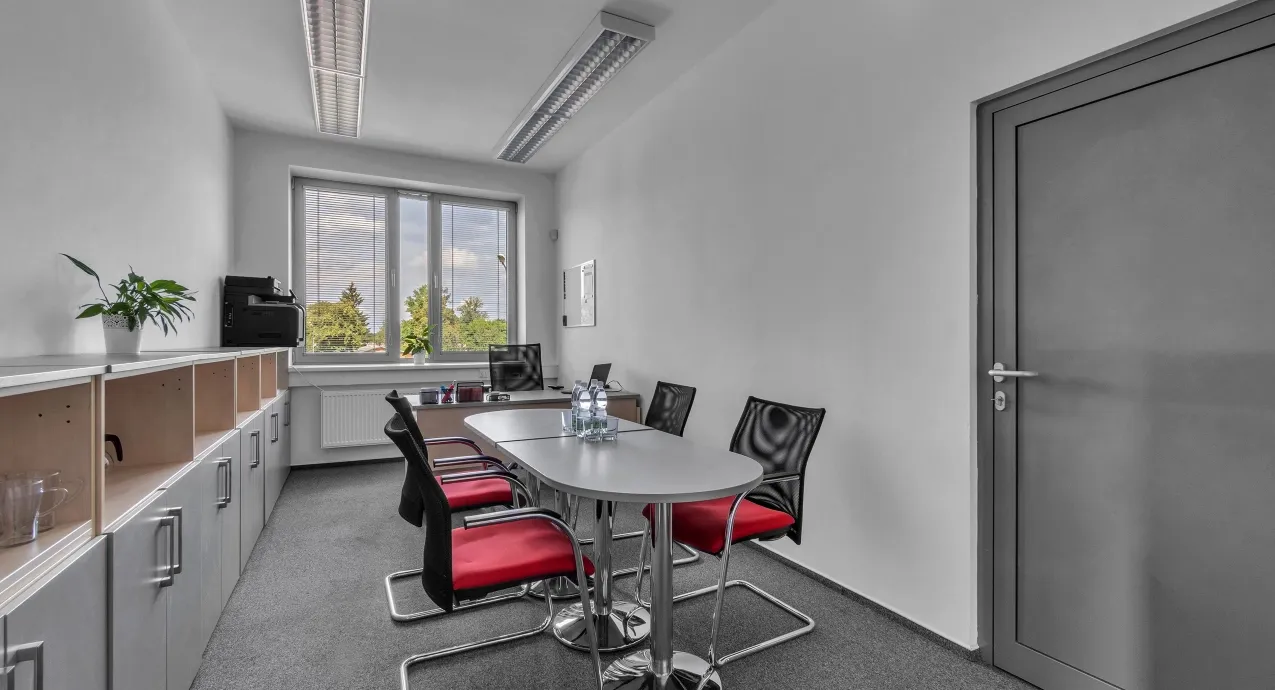 Modern meeting room with oval table, red and black chairs, large window, and storage cabinets.