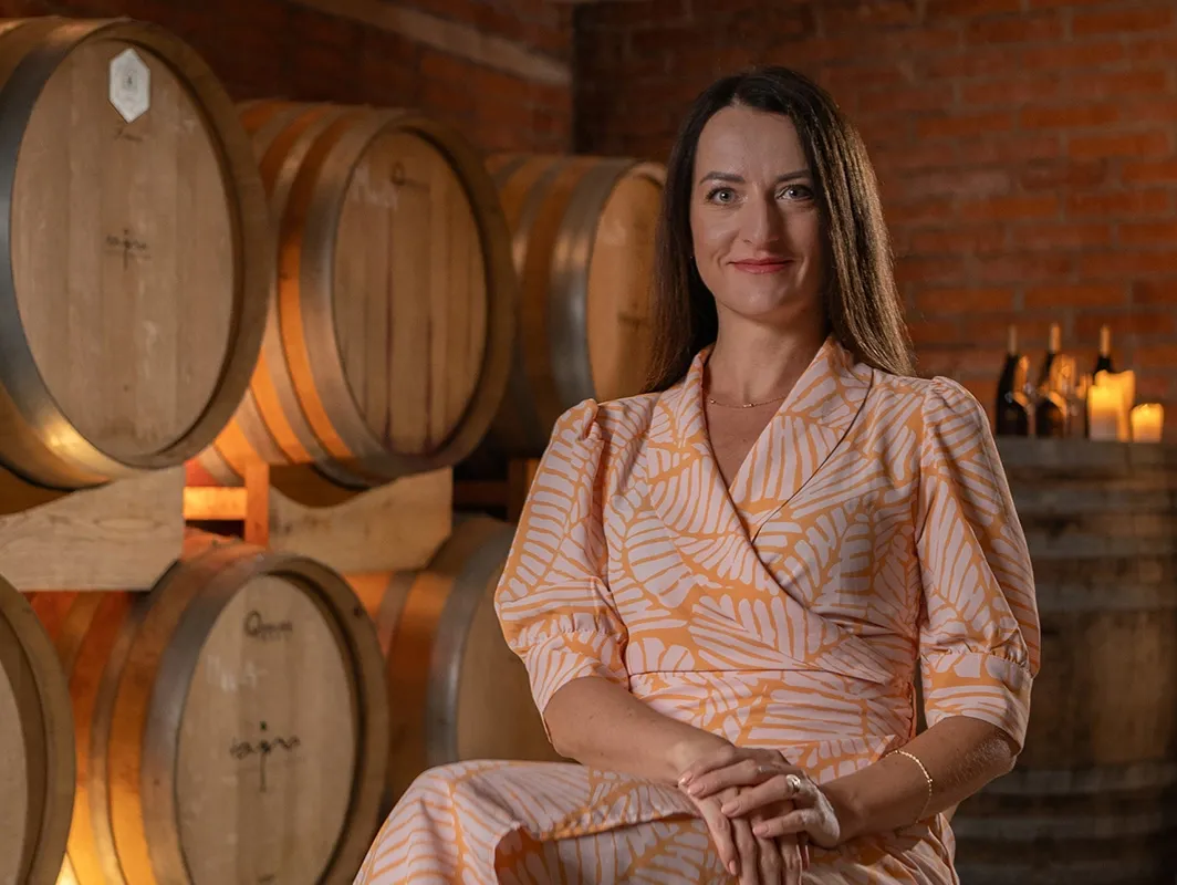 Woman in a patterned dress sits among wine barrels in a warm cellar.